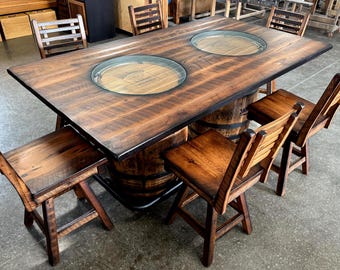 May include: A rectangular wooden table with a dark brown finish, supported by two whiskey barrels. The tabletop features two circular glass insets, one displaying the Jack Daniel's logo. Four matching wooden bar stools with slatted backrests.