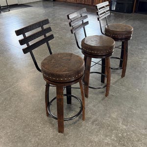 May include: Three swivel bar stools featuring dark brown, textured, circular seats with decorative metal nailhead trim. Each stool has a curved backrest made of three horizontal wooden slats and a black metal frame with a circular footrest.