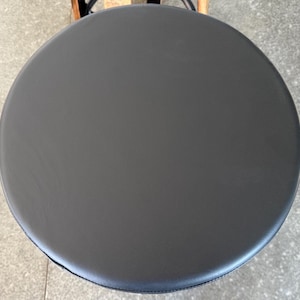 May include: A circular stool seat featuring a smooth, matte black surface. The top is perfectly round with a clean, uniform finish, showing no visible textures or patterns. The edges are slightly rounded, creating a simple, functional design.