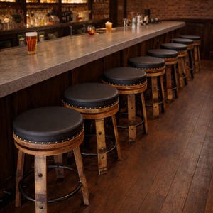 May include: A bar featuring a row of black leather-topped stools with wooden legs and metal footrests. The bar has a concrete countertop and a brick wall backdrop. A pint of beer sits on the bar.
