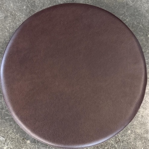 May include: A circular stool seat cushion with a dark brown, textured faux leather surface. The cushion features a smooth, rounded edge and a consistent, matte finish across the entire top, providing a simple and functional design element.