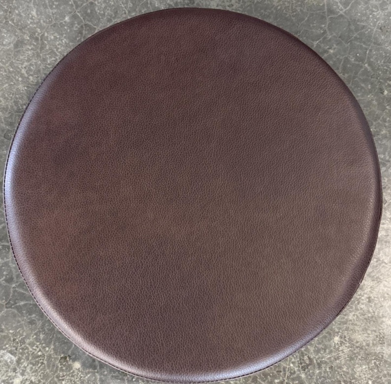 May include: A circular stool seat cushion with a dark brown, textured faux leather surface. The cushion features a smooth, rounded edge and a consistent, matte finish across the entire top, providing a simple and functional design element.