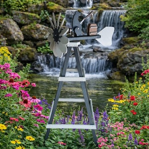 Puede incluir: Un molino de viento decorativo de madera con un oso pescando en la parte superior, sobre un fondo de cascada y exuberante jardín. El molino de viento presenta una combinación de colores gris y blanco con una veleta en forma de flor.