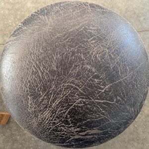 May include: A circular stool seat featuring a dark brown, textured surface with a prominent, weathered, and cracked faux leather pattern. The material displays a complex network of fine lines and creases across the entire round top surface.