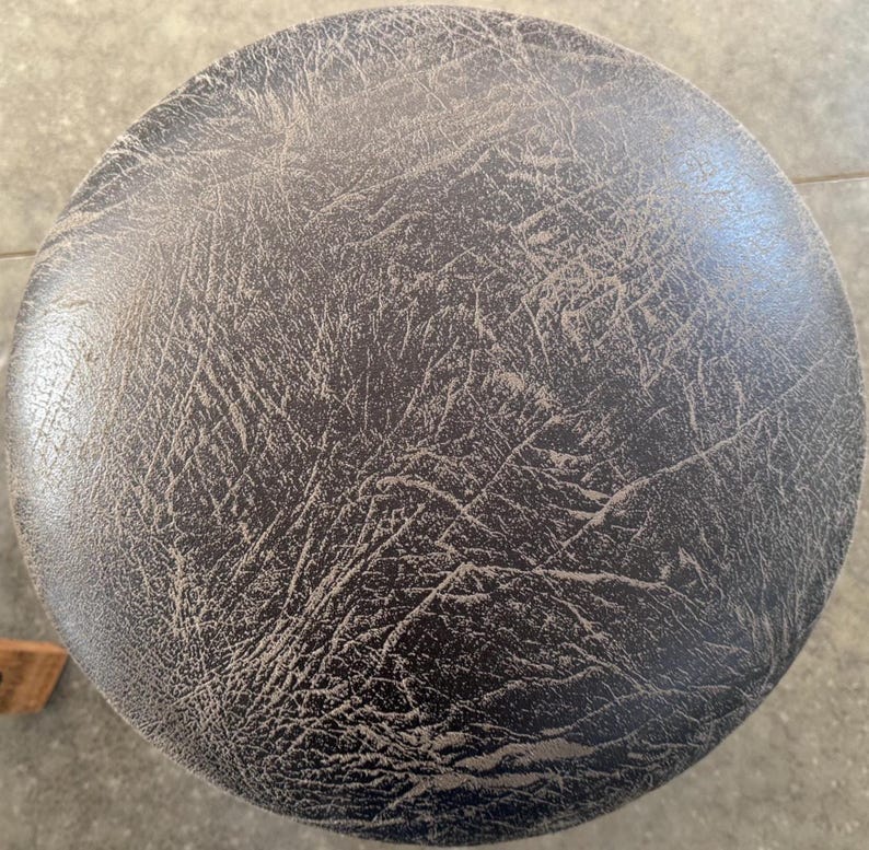 May include: A circular stool seat featuring a dark brown, textured surface with a prominent, weathered, and cracked faux leather pattern. The material displays a complex network of fine lines and creases across the entire round top surface.