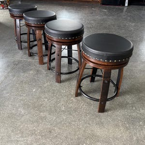 May include: Four swivel bar stools featuring dark brown wooden barrel-style legs and circular black padded seats. Each stool includes a black metal footrest ring and a row of silver-toned decorative nailhead trim around the edge of the seat cushion.