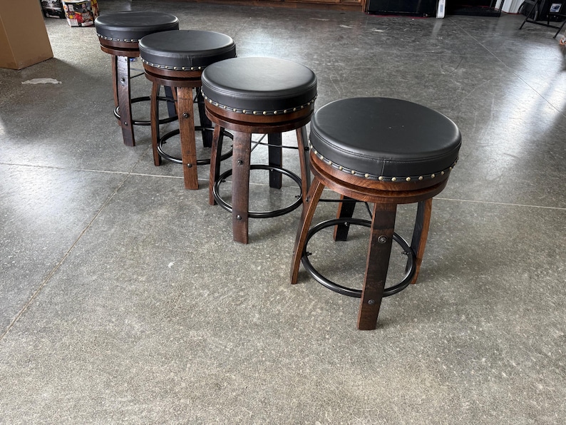 May include: Four swivel bar stools featuring dark brown wooden barrel-style legs and circular black padded seats. Each stool includes a black metal footrest ring and a row of silver-toned decorative nailhead trim around the edge of the seat cushion.