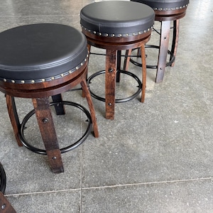 May include: A set of three bar stools featuring dark brown wooden legs with a curved design, a circular black padded seat, and a black metal footrest ring. The edge of each seat is accented with a row of silver-toned metal nailhead trim studs.