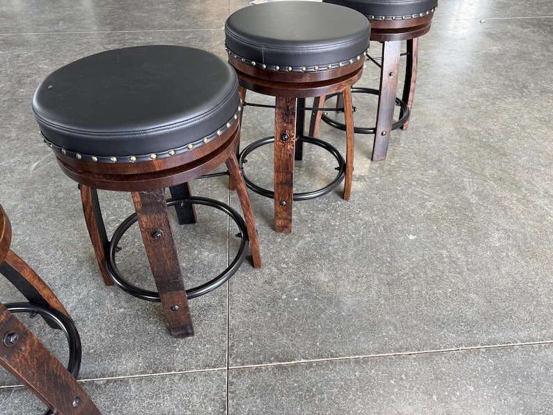 May include: A set of three bar stools featuring dark brown wooden legs with a curved design, a circular black padded seat, and a black metal footrest ring. The edge of each seat is accented with a row of silver-toned metal nailhead trim studs.
