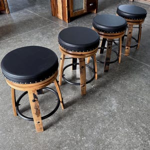 May include: Three round stools featuring black padded seats with silver nailhead trim. The bases are constructed from curved wooden barrel staves with a natural finish, reinforced by black circular metal footrests connecting the legs.