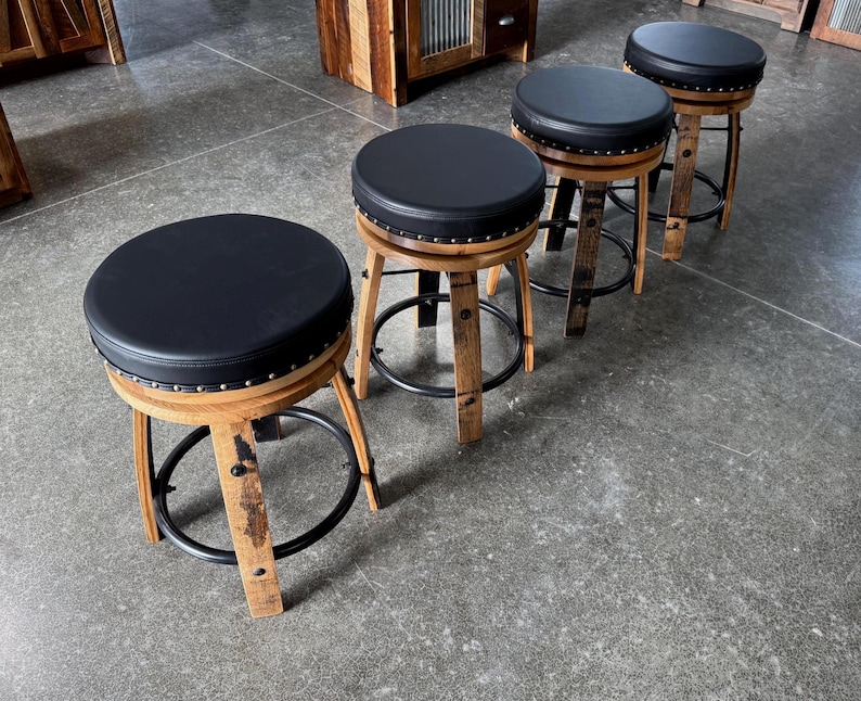 May include: Three round stools featuring black padded seats with silver nailhead trim. The bases are constructed from curved wooden barrel staves with a natural finish, reinforced by black circular metal footrests connecting the legs.