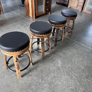 May include: A set of four bar stools featuring round black padded seats with silver nailhead trim. The bases are constructed from curved light brown wood staves with a circular black metal footrest connecting the legs of each stool.