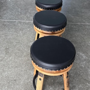 May include: A row of four round bar stools featuring black padded circular seats with silver nailhead trim. The base of each stool is constructed from curved light brown wood staves, resembling a barrel, and includes a black metal foot ring.