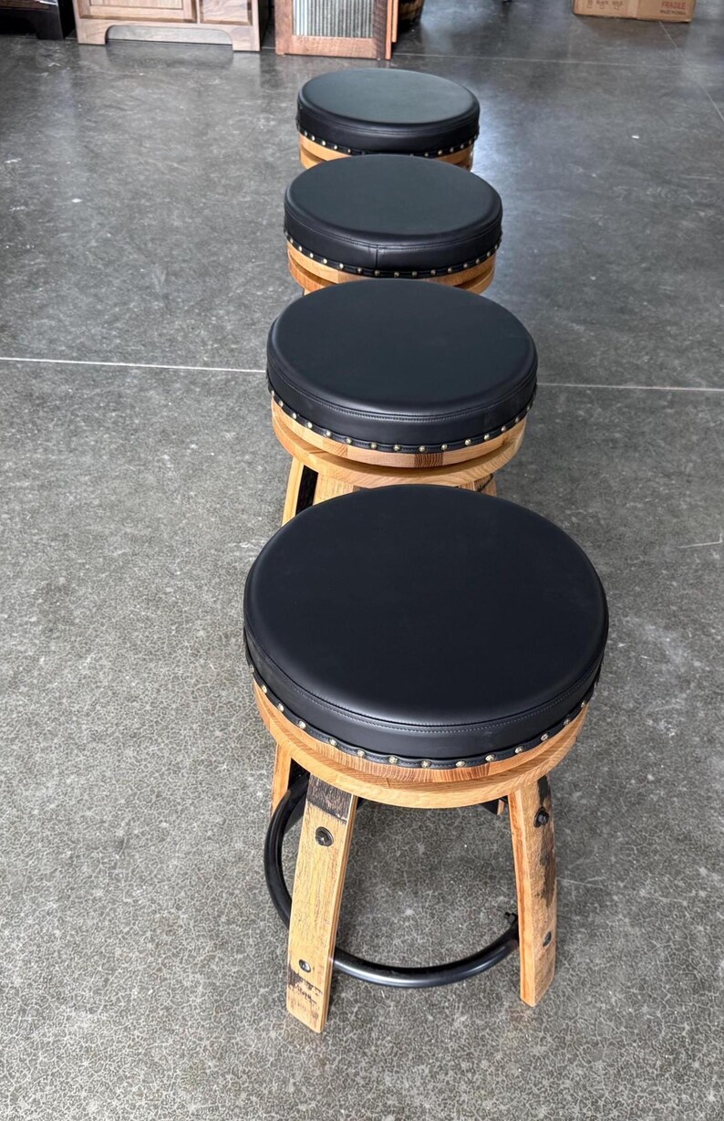 May include: A row of four round bar stools featuring black padded circular seats with silver nailhead trim. The base of each stool is constructed from curved light brown wood staves, resembling a barrel, and includes a black metal foot ring.