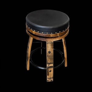 May include: A round stool featuring a black padded seat with brass nailhead trim. The base is constructed from curved wooden staves with a natural finish, reinforced by a circular black metal footrest ring that connects the four legs.