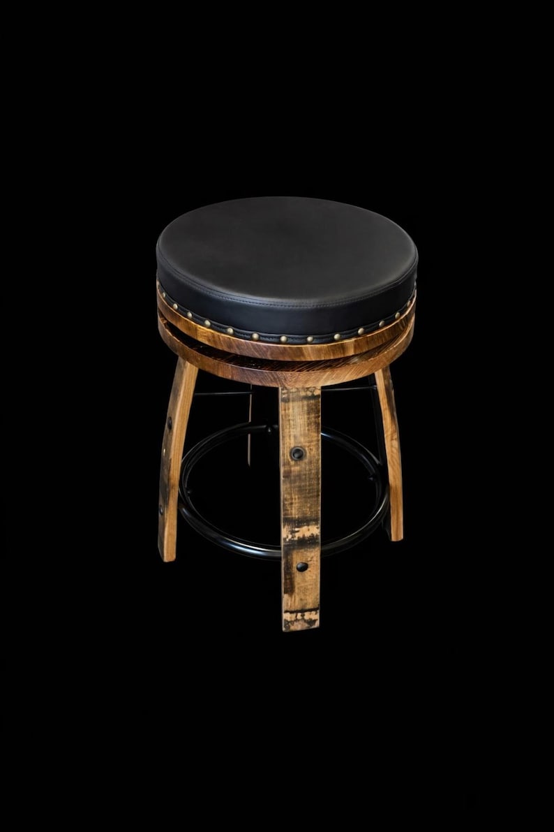 May include: A round stool featuring a black padded seat with brass nailhead trim. The base is constructed from curved wooden staves with a natural finish, reinforced by a circular black metal footrest ring that connects the four legs.