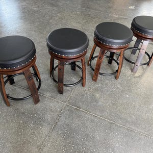 May include: Four round stools with black padded seats and silver nailhead trim. Each stool features curved dark brown wooden legs connected by a circular black metal footrest ring. The stools are arranged in a row on a grey floor surface.