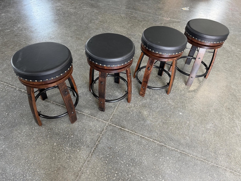May include: Four round stools with black padded seats and silver nailhead trim. Each stool features curved dark brown wooden legs connected by a circular black metal footrest ring. The stools are arranged in a row on a grey floor surface.