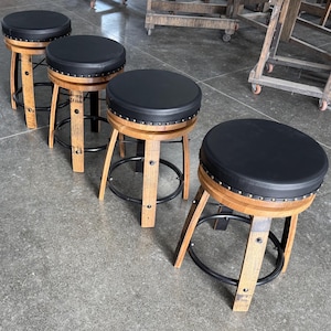 May include: Four backless bar stools featuring circular black padded seats with silver nailhead trim. The bases are constructed from curved wooden barrel staves with black metal circular footrests connecting the legs for structural support.