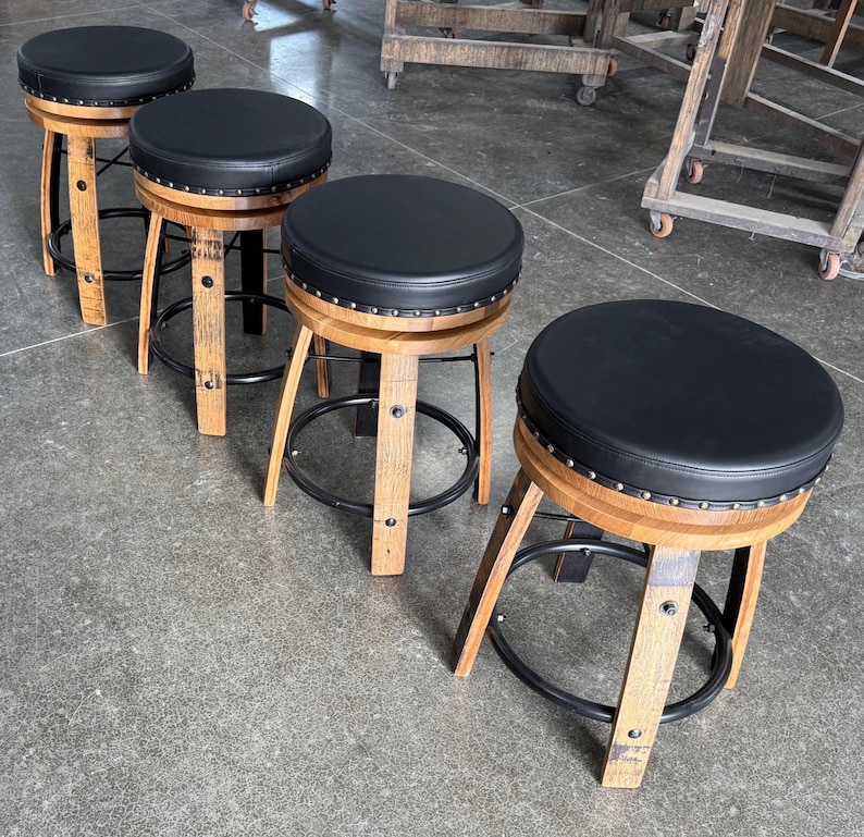 May include: Four backless bar stools featuring circular black padded seats with silver nailhead trim. The bases are constructed from curved wooden barrel staves with black metal circular footrests connecting the legs for structural support.