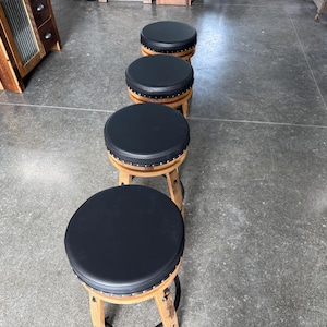 May include: A row of four round stools with black padded seats and wooden bases. Each stool features a circular top upholstered in smooth black material, accented with a row of small metal studs along the edge, supported by curved wooden legs.