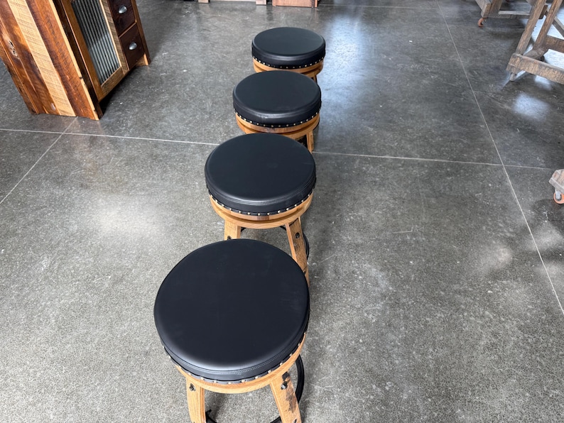 May include: A row of four round stools with black padded seats and wooden bases. Each stool features a circular top upholstered in smooth black material, accented with a row of small metal studs along the edge, supported by curved wooden legs.