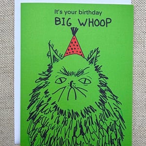 May include: A green birthday card with the text "It's your birthday BIG WHOOP." The card features a cartoon cat wearing a red and white party hat. The cat has a grumpy expression and is drawn in black ink.
