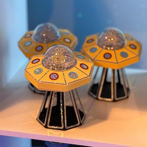 May include: Three yellow and black UFO-shaped decorations with clear domes filled with glitter. The UFOs have colorful circular accents and sit on a white surface. The decorations have a retro, space-themed aesthetic.