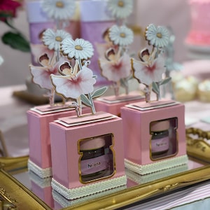 May include: Pink gift boxes with a ballerina and daisy topper, each containing a small jar with the name "Helena" on a label. The boxes are on a gold tray, with a blurred background of more boxes and a cake.
