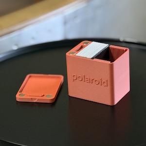 Pode incluir: Um porta-filmes Polaroid cor coral com uma tampa a condizer. O suporte tem formato de cubo com a palavra "Polaroid" em relevo na frente. A tampa tem dois pequenos ímanes circulares. O suporte contém uma pilha de filmes.