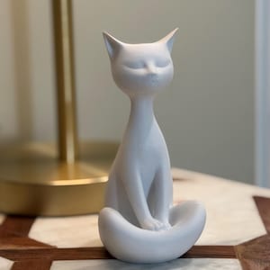 May include: A white ceramic cat figurine in a seated pose with its eyes closed. The cat has a slender neck and a curved base, creating a minimalist and zen-like aesthetic. The figurine is placed on a marble surface.