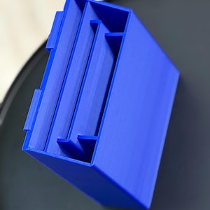 May include: A bright blue 3D-printed maze game. The rectangular puzzle has a series of internal walls creating a complex path. The top edge features small tabs. The object is set against a dark background.