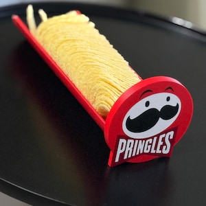 May include: A red Pringles-branded chip holder filled with golden potato chips. The holder features the Pringles logo with a mustache and the word "PRINGLES" in white text. The chips are stacked neatly.