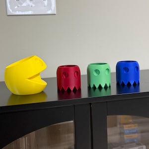 May include: A yellow Pac-Man-shaped container and three ghost-shaped containers in red, green, and blue. The containers have jagged edges at the bottom and circular eye cutouts. The yellow container has an open mouth.
