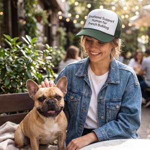May include: A trucker hat with a white front panel and a dark green brim and mesh back. The front panel features the black printed text: Emotional Support Human for French Bulldog. The hat has a structured crown and an adjustable snapback closure.