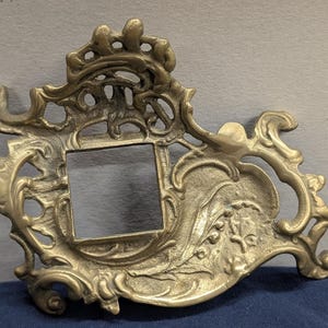 May include: Ornate, antique-style metal frame with a square opening. The frame has a detailed, scrolling design with floral and foliate elements. The color is a muted gold or brass tone, suggesting an aged finish. The frame is likely for a picture or mirror.