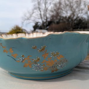 Könnte beinhalten: Eine Vintage-Schale aus türkisfarbener Keramik mit gewelltem Rand und goldenem Besatz. Die Schale hat ein Blumendesign in Gold und Weiß. Die Schale steht auf einer weißen Oberfläche, mit einem verschwommenen Hintergrund im Freien.