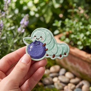 May include: An embroidered patch featuring a light mint green caterpillar with a smiling face, holding a large, round, dark purple blueberry with a small green leaf attached to the side. The patch has a stitched border and brown feet.