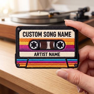 May include: A rectangular embroidered patch shaped like a vintage cassette tape. The design features horizontal stripes in orange, yellow, and purple, with white labels reading CUSTOM SONG NAME and ARTIST NAME in black text and two black spools.