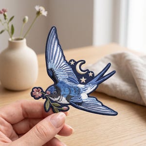 May include: An embroidered patch featuring a blue swallow in flight, holding a small pink flower with green leaves in its beak. The design includes a white crescent moon and three small stars against a dark blue background, with detailed stitching.