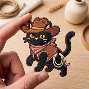 May include: An embroidered patch featuring a black cat wearing a brown cowboy hat, a brown vest, and a pink bandana around its neck. The cat has yellow eyes and is holding a coiled white lasso rope in its tail, rendered in detailed stitching.