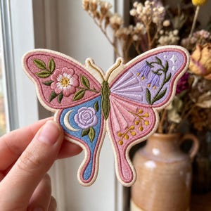May include: An embroidered butterfly patch featuring a split design. The left wing displays a pink flower and a purple rose with a crescent moon on a blue background. The right wing shows purple bell-shaped flowers and golden sprigs on a pink base.