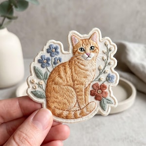 May include: An embroidered patch featuring a seated orange tabby cat surrounded by blue and rust-colored flowers and green leaves. The design is stitched onto a cream-colored fabric base with a scalloped edge, showcasing detailed thread work.