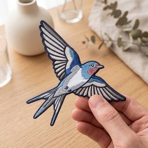 May include: An embroidered swallow patch featuring a bird in flight with wings spread wide. The design incorporates shades of blue, white, grey, and a touch of reddish-pink on the throat, all outlined with a dark navy blue border for definition.