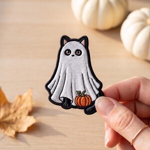 May include: An embroidered patch featuring a black cat wearing a white ghost costume. The cat has large dark eyes and is holding a small orange pumpkin with a green stem. The patch has a thick black border and detailed white stitching.