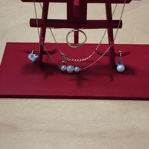 以下が含まれることがあります： 日本の鳥居のデザインの赤い木製ディスプレイスタンド。ゴールドの指輪、パールネックレス、パールイヤリングなどのジュエリーを展示しています。スタンドは薄茶色の表面にあります。