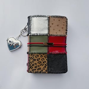 Puede incluir: Una cubierta de diario rectangular con un diseño de patchwork de varias texturas y colores, incluyendo plata, beige, verde oliva, rojo, estampado de leopardo y negro. Está unida por costuras negras y asegurada con bandas elásticas rojas.