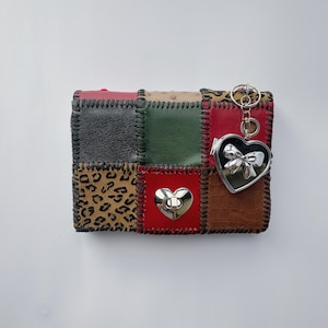 May include: A rectangular wallet featuring a patchwork design of red, black, green, brown, and leopard print panels joined by dark stitching. It includes a silver heart-shaped turn-lock closure and a dangling silver heart charm with a bow detail.