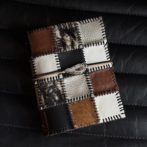 Puede incluir: Una cubierta de diario rectangular con un diseño de patchwork de cuero marrón con relieve de cocodrilo, piel de vaca en blanco y negro, cuero plateado metalizado y cuadrados de cuero negro sólido, unidos por costuras negras gruesas.