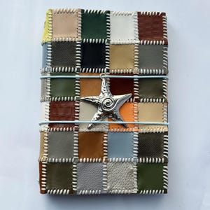 Könnte beinhalten: Ein rechteckiger Notizbuchumschlag mit einem Patchwork-Design aus verschiedenfarbigen Lederquadraten in Braun-, Grün-, Grau-, Weiß- und Gelbtönen. Die Quadrate sind mit weißen Überwendlichstichen verbunden und ein silberner Seestern-Anhänger ist befestigt.
